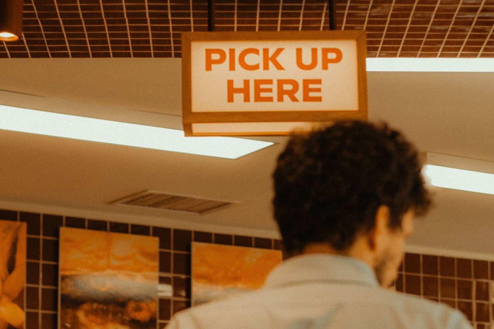 A customer waits beneath a lit "Pick Up Here" sign at a restaurant takeout counter